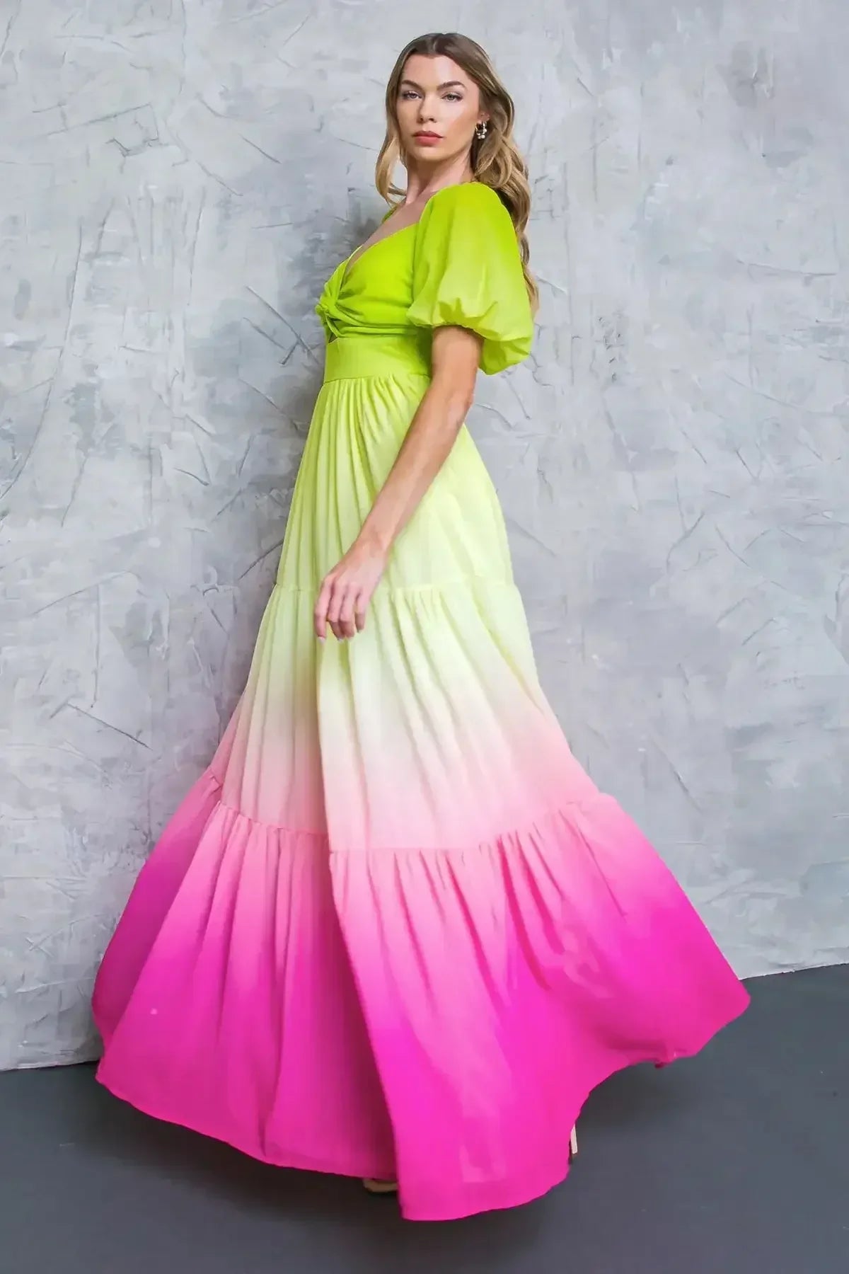 A model wears a vibrant, gradient dress transitioning from green to pink, elegantly showcasing its flowy design against a textured gray background. The setting enhances the dress's colorful aesthetic.