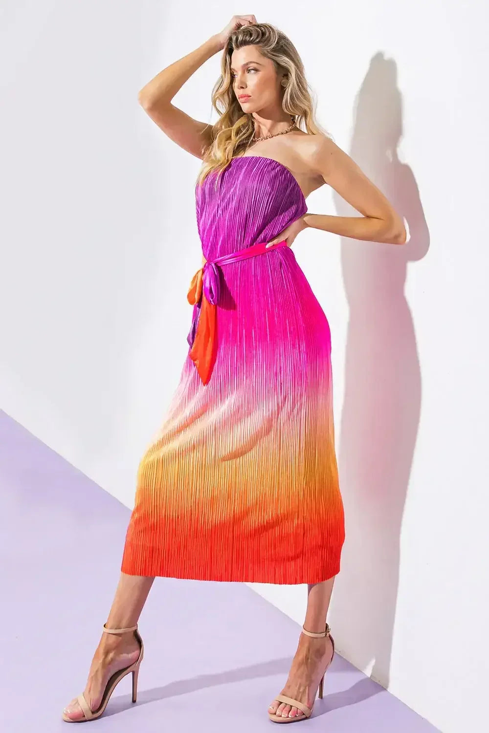 A woman poses confidently in a vibrant, pleated, strapless dress that transitions from purple to orange. She stands against a white wall, with a pastel purple floor beneath her.