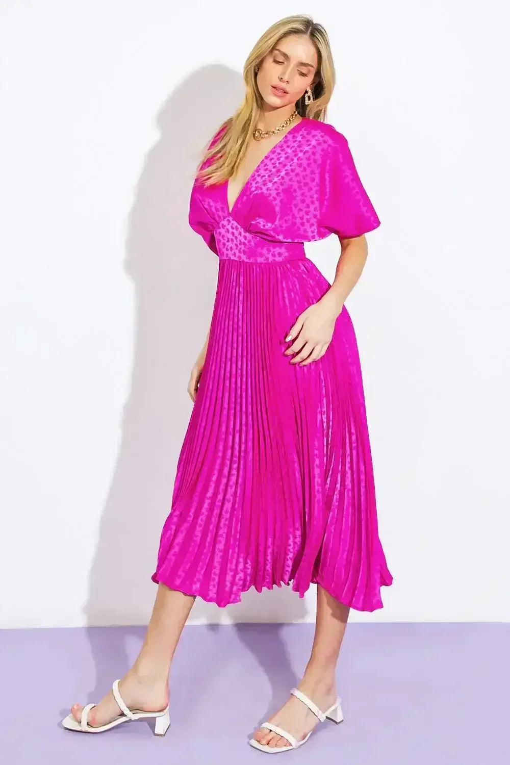 A woman stands gracefully in a vivid pink, pleated dress. She poses with one hand on her hip, complemented by white strappy heels, against a minimalist white and purple backdrop.