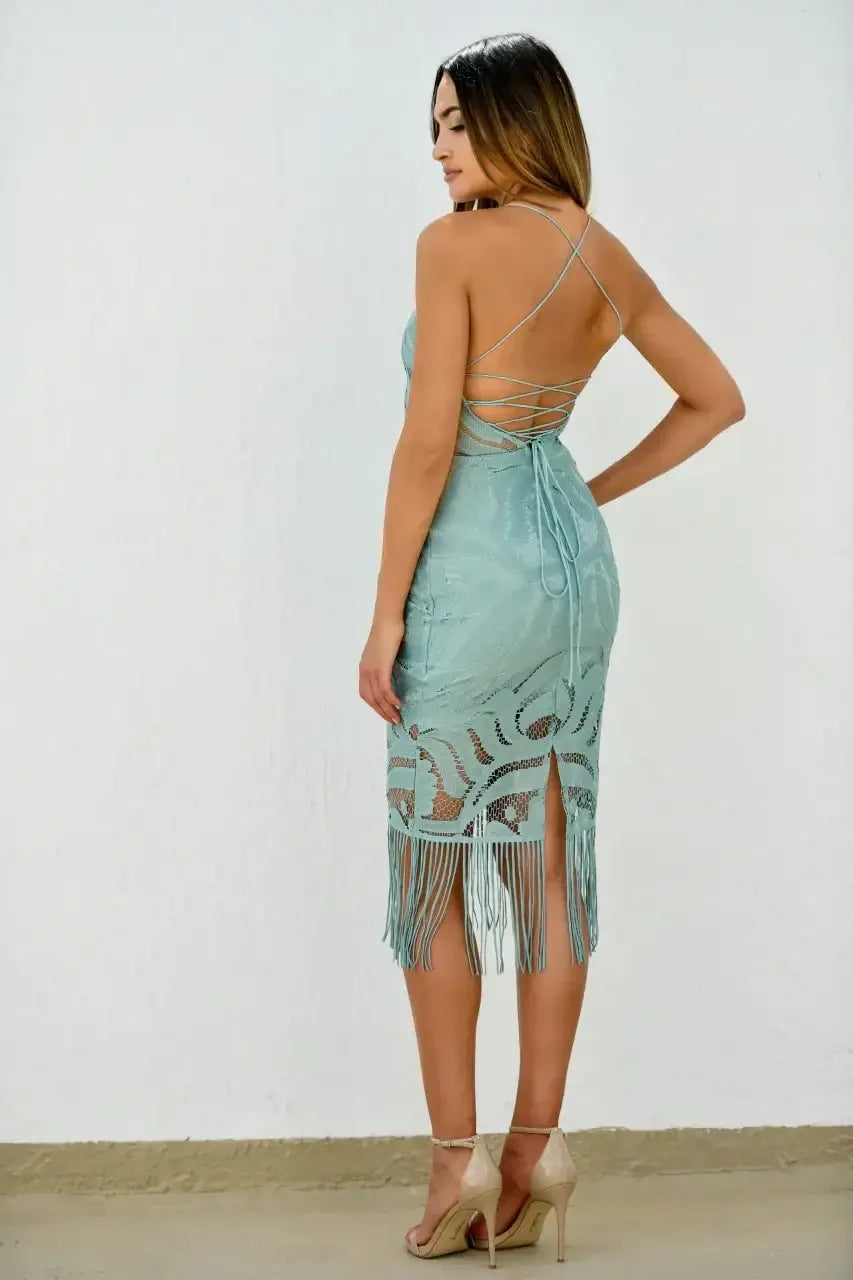 A woman stands facing away, wearing a light blue dress with intricate lace patterns and fringe at the hem. She poses against a plain white wall, showcasing the dress's back design.