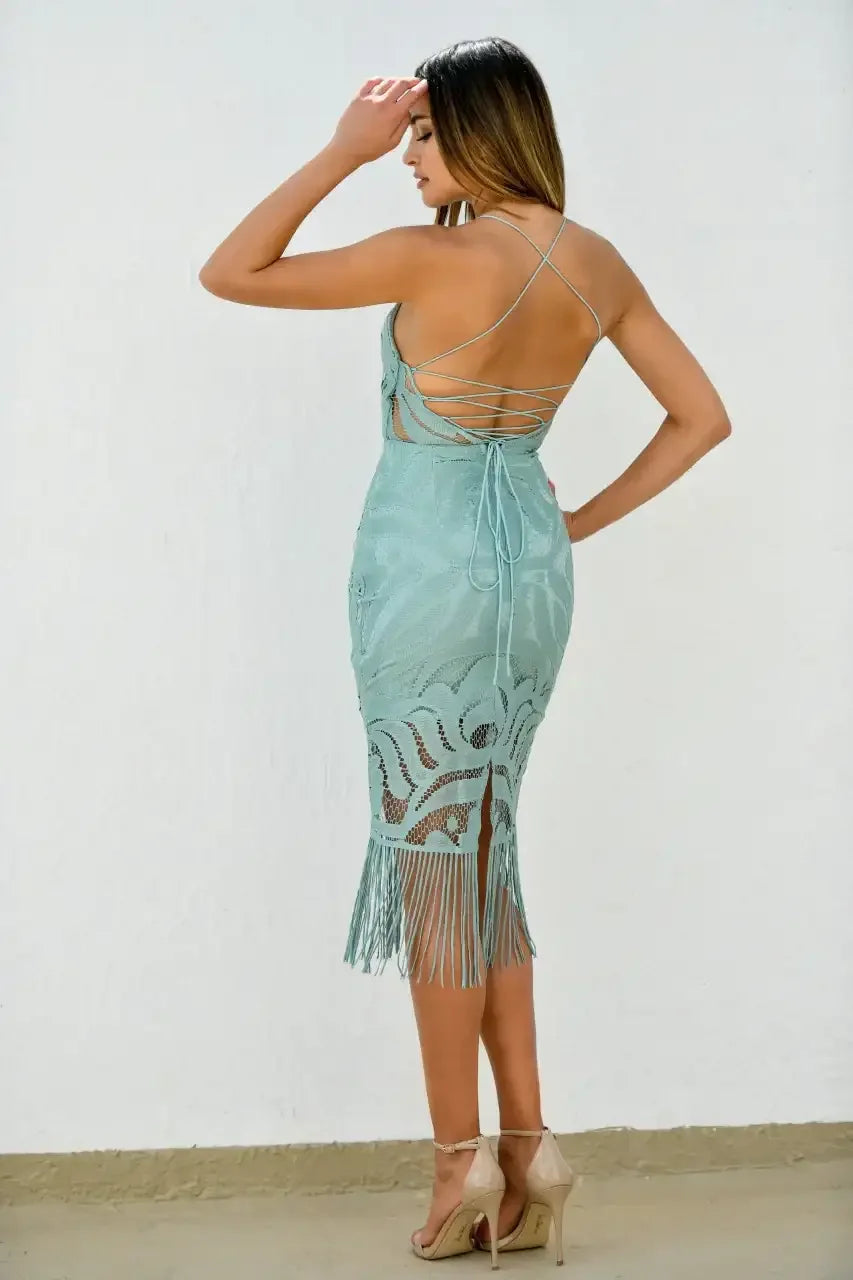 A woman stands with her back to the viewer, wearing a teal dress with intricate lace and fringes. She gazes to the side against a textured white wall and beige floor.