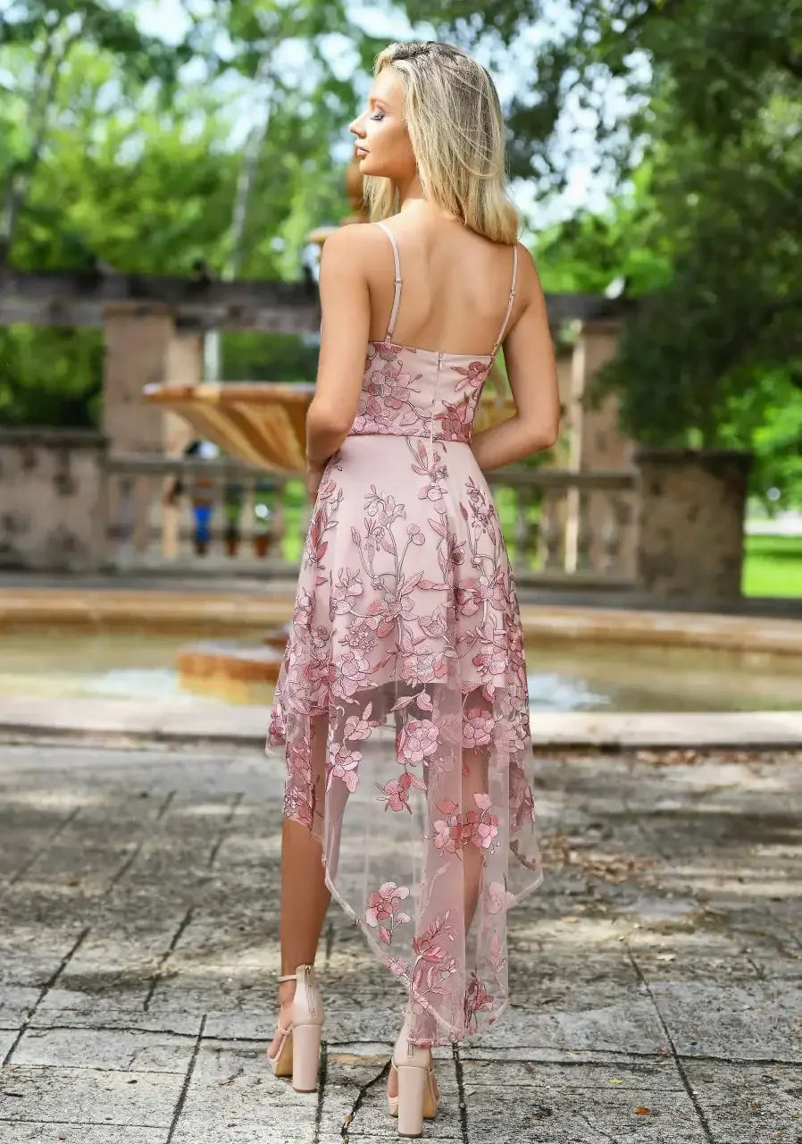 A woman stands gracefully in a pink floral dress, facing away from the camera, in a lush garden with a fountain in the background on a stone-paved surface.