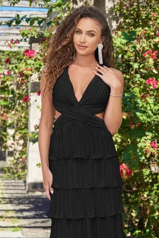 A woman stands confidently in a layered black dress, showcasing her curly hair and accessories. She poses amidst a backdrop of lush greenery and vibrant flowers, suggesting a serene outdoor setting.