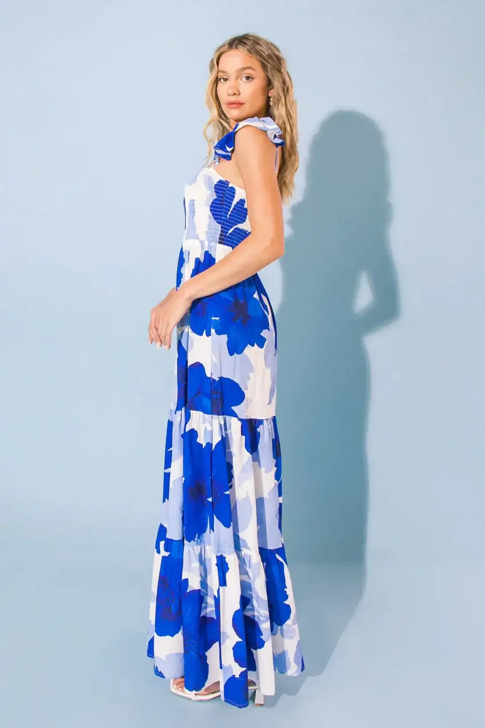 A woman stands sideways, showcasing a long, floral dress with blue and white patterns. She poses against a light blue background, her hair cascading in loose waves.
