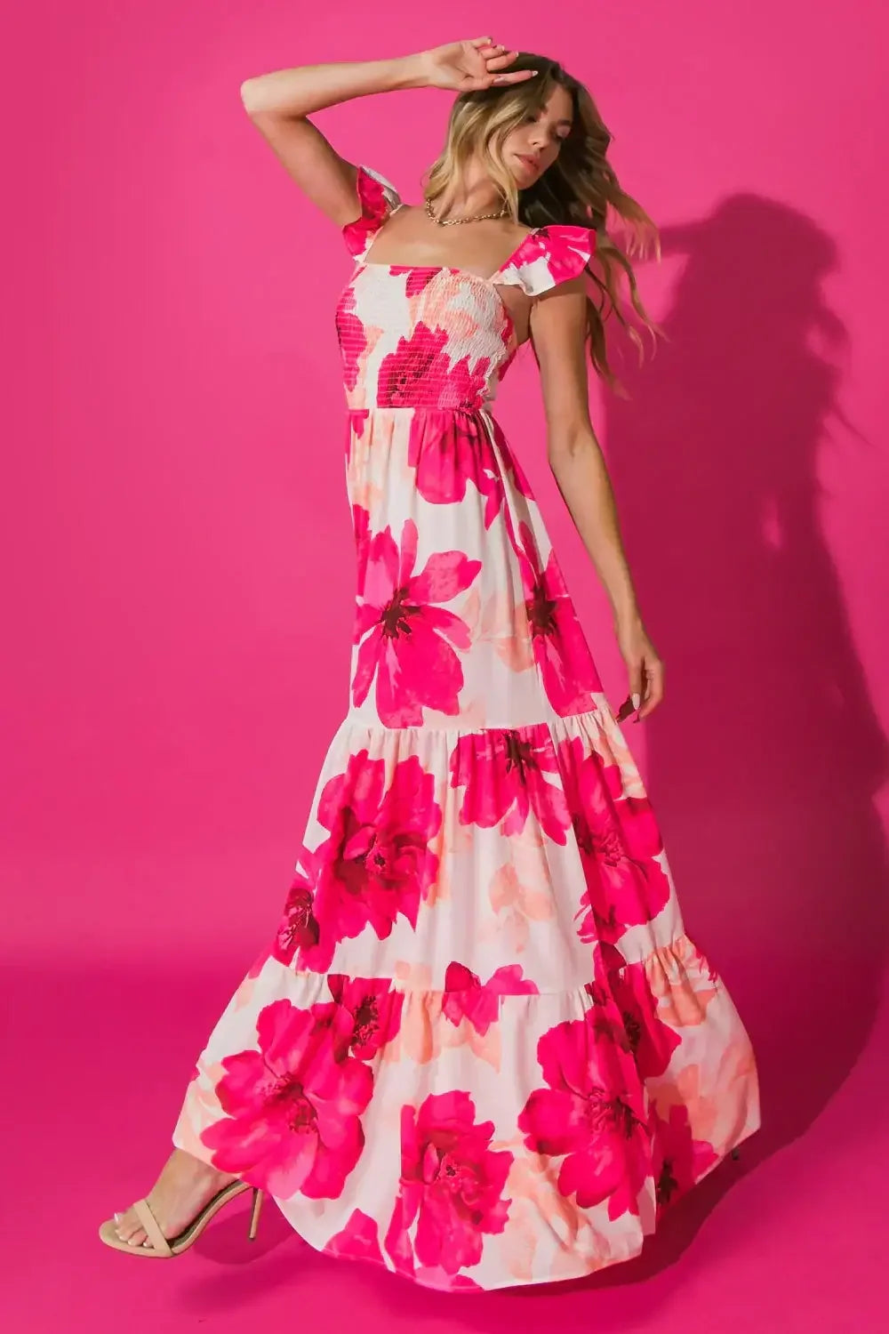 A woman models a long, floral dress with pink flowers, dancing gracefully against a bright pink backdrop, highlighting the dress's movement and vibrant colors.