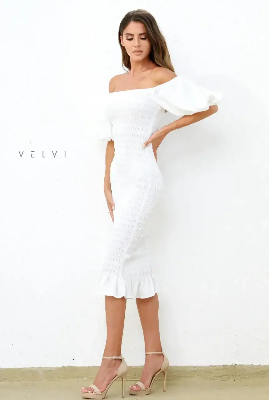 A woman wearing a fitted white off-shoulder dress with puff sleeves stands gracefully against a textured white wall. She pairs the outfit with neutral high-heeled sandals, exuding elegance.
