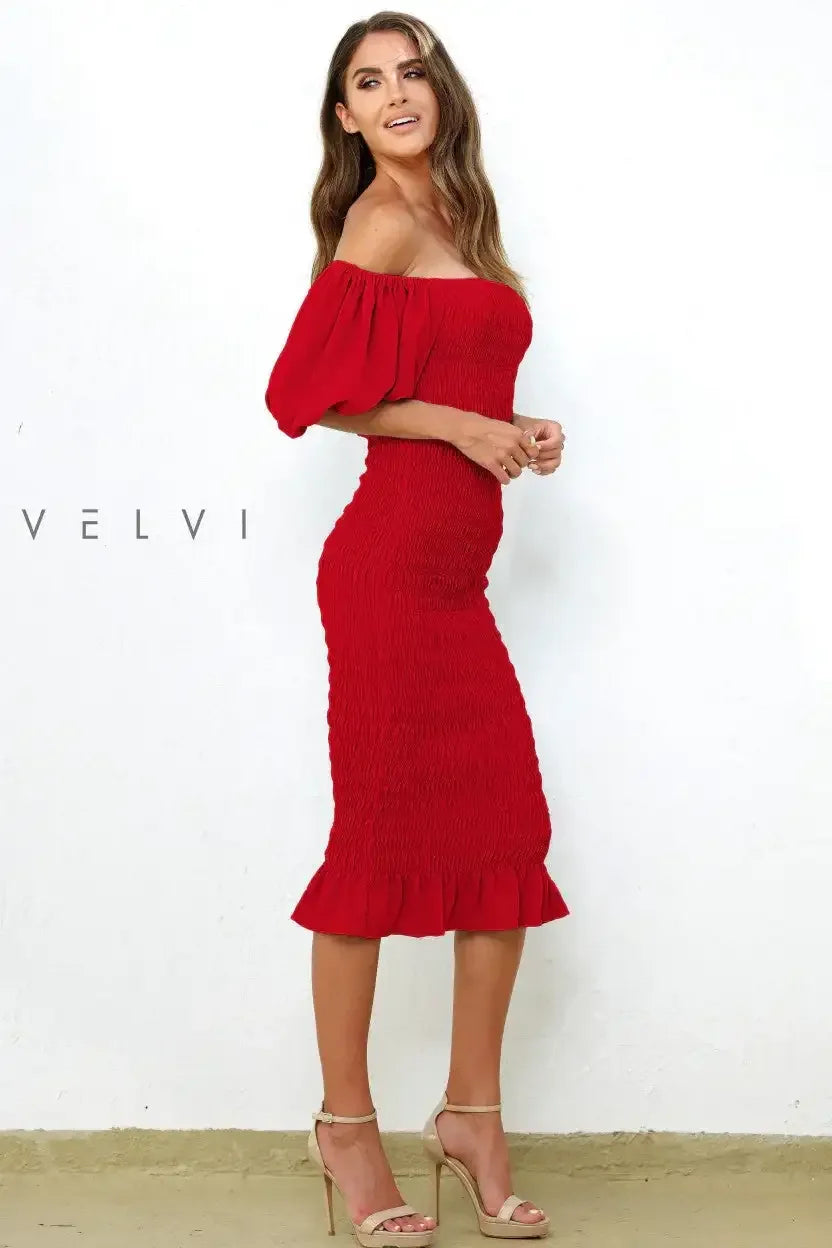 A woman in a fitted red dress with off-the-shoulder sleeves stands against a white wall, smiling gracefully. She's wearing strappy beige heels, showcasing an elegant pose. Text: "V?LV?."