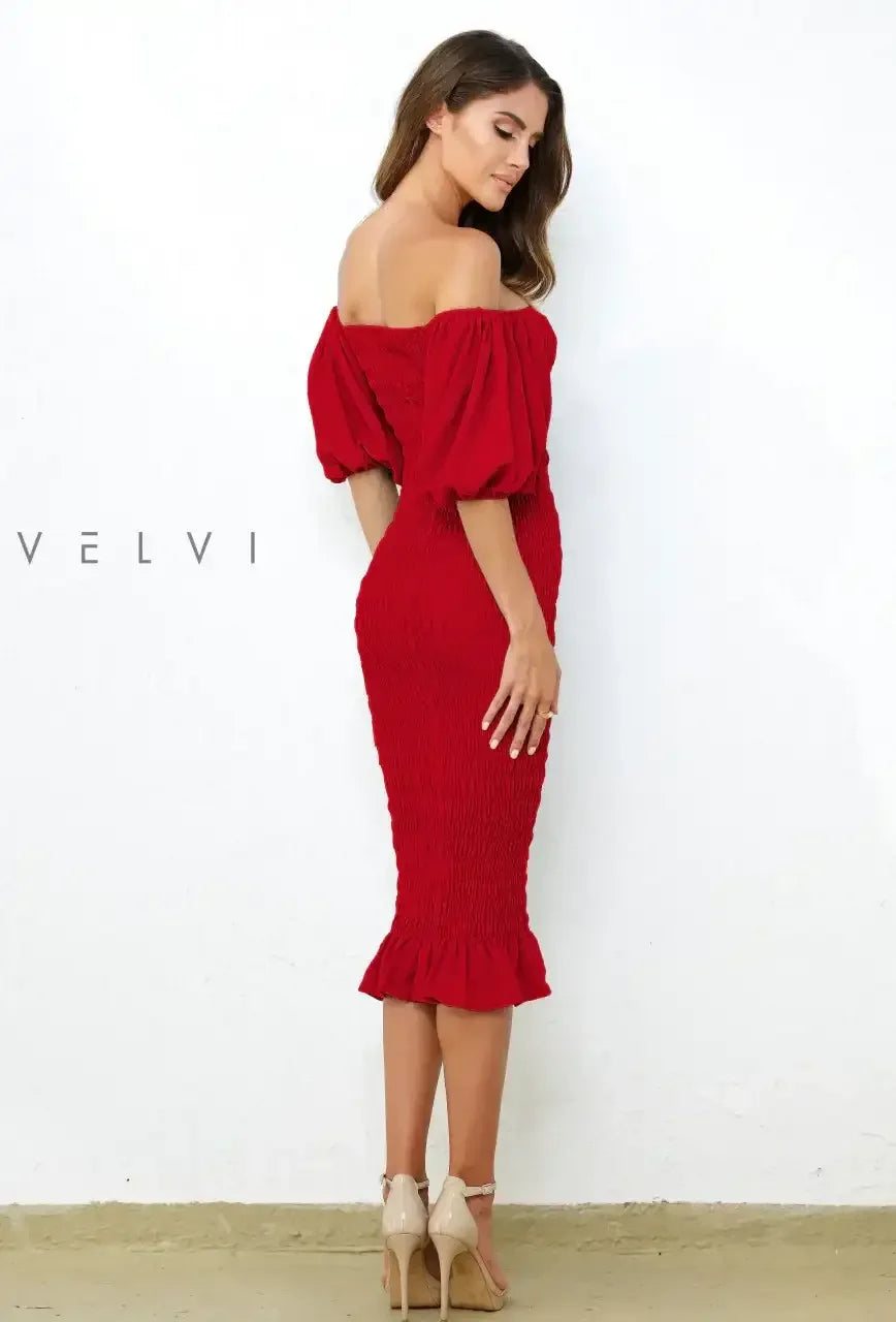 A woman in a red off-the-shoulder dress stands elegantly, showcasing gathered fabric and a flared hem. She faces away against a white wall, while her hair cascades softly down her back.