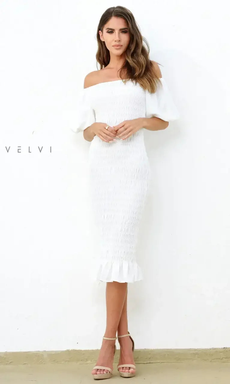 A woman stands confidently, wearing a fitted white off-the-shoulder dress with a ruffled hem, against a textured white wall and light-colored floor, displaying elegance and style.