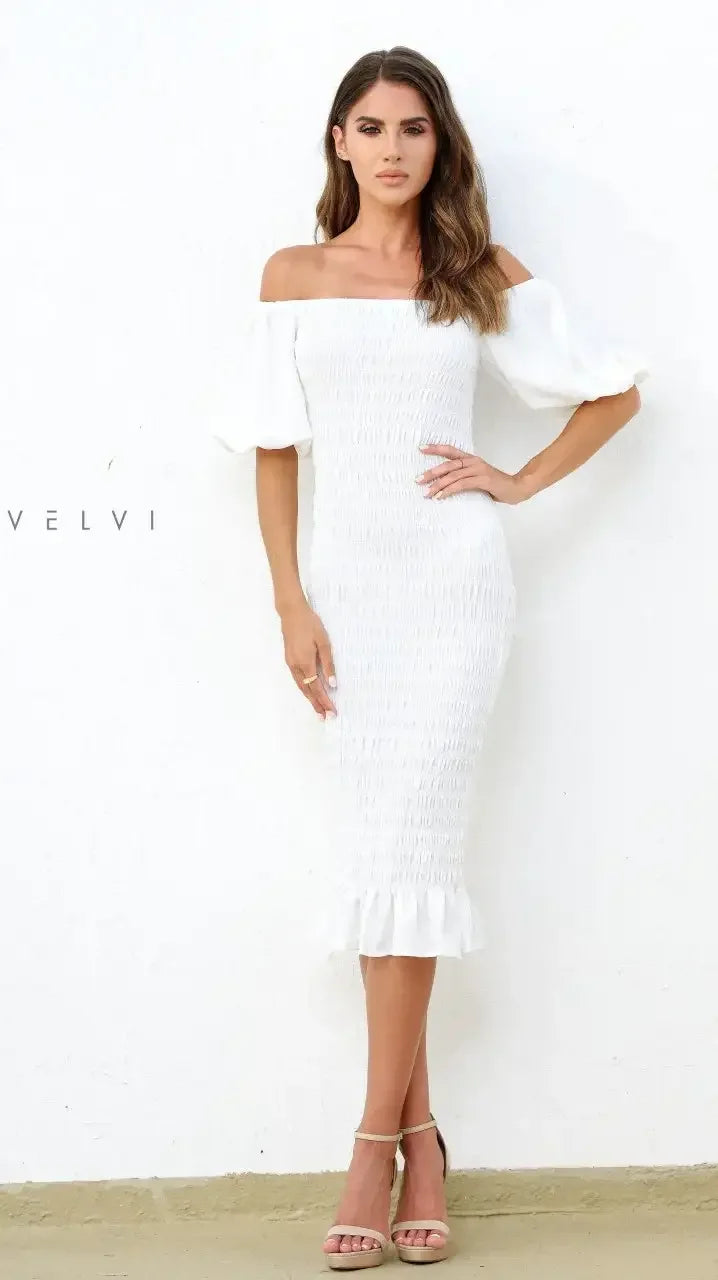 A woman stands confidently in a fitted, off-the-shoulder white dress with puff sleeves, showcasing a textured pattern. She poses against a plain white wall, wearing neutral sandals.