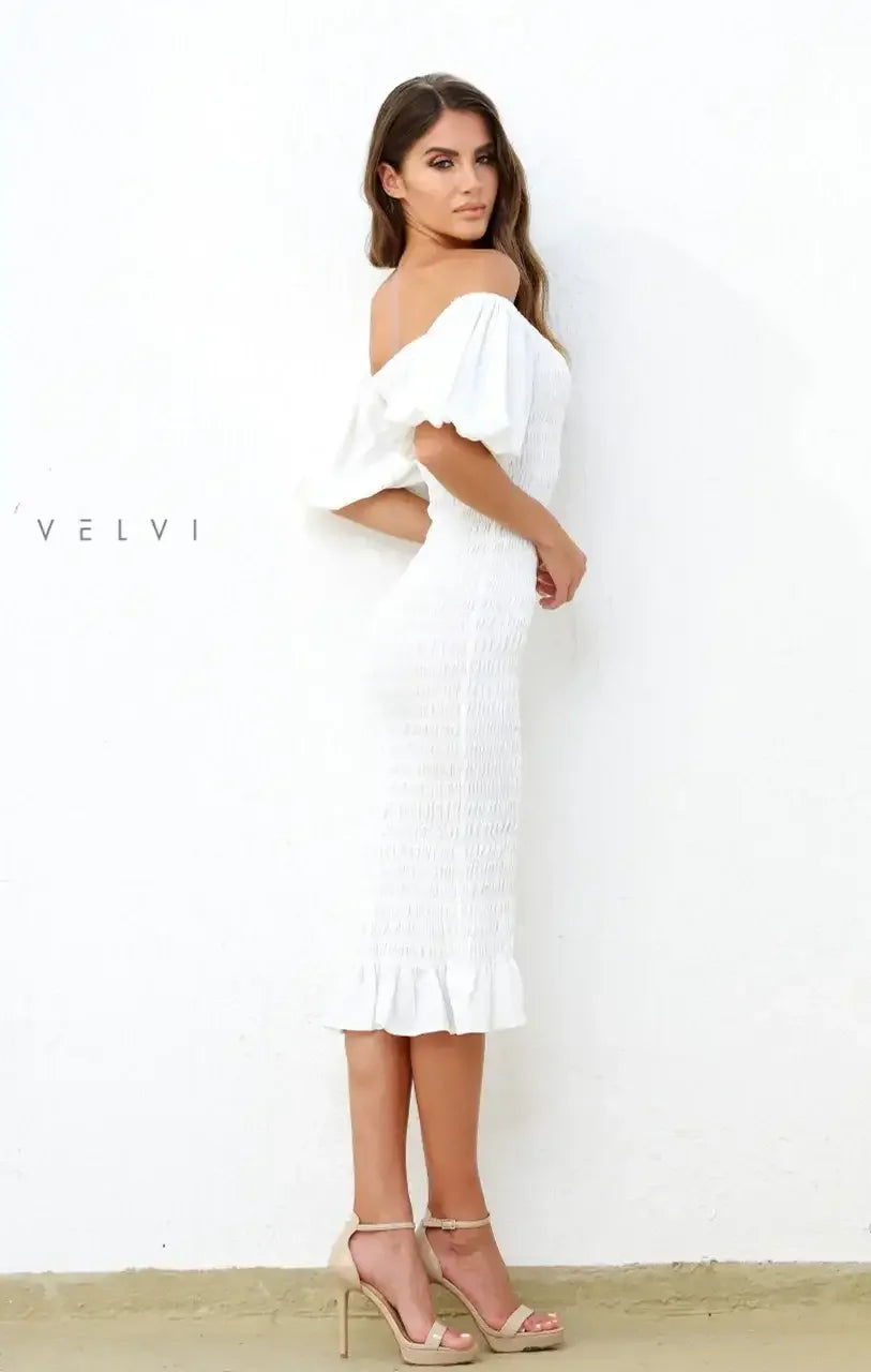 A woman in a form-fitting white dress with puff sleeves stands against a plain white wall. She poses elegantly, showcasing her outfit and wearing light-colored high heels.
