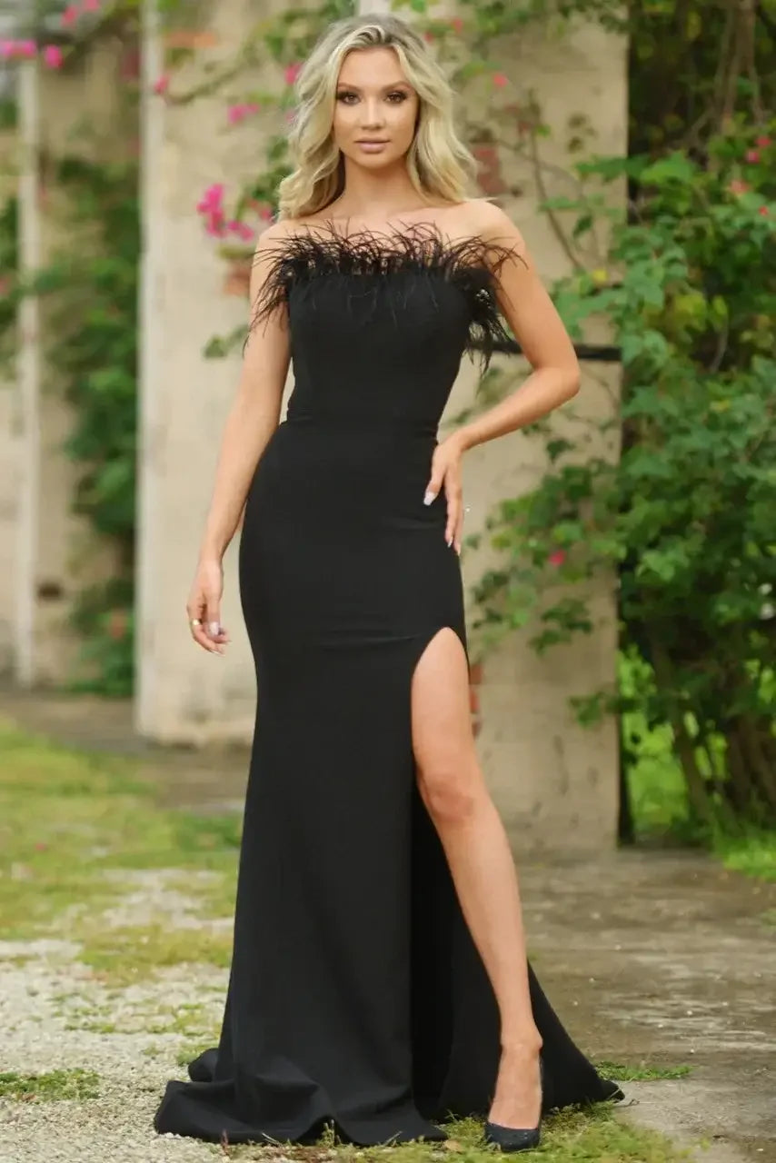 A woman stands confidently in a black, form-fitting dress with feathered detailing at the neckline and a thigh-high slit, surrounded by lush greenery and concrete structures.