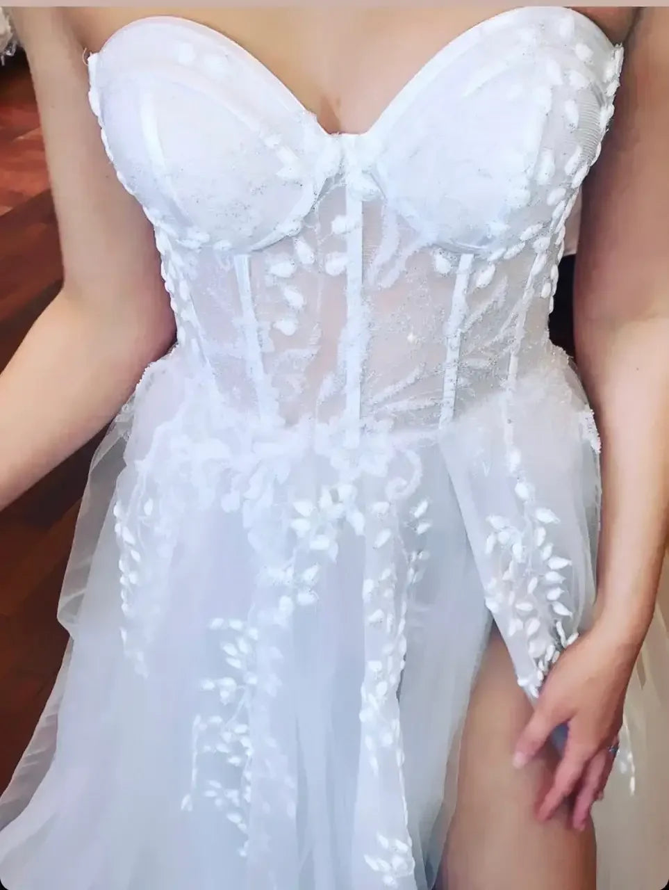 A white strapless dress features intricate floral embroidery and a sheer skirt. The wearer adjusts the dress, with a focus on the elegant details set against a warm wooden floor.