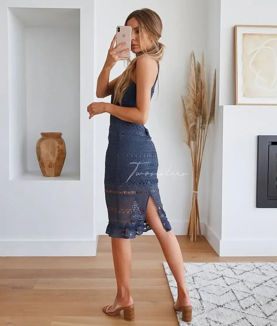 A woman poses while taking a selfie, wearing a navy lace dress with a side slit. The setting includes a minimalist interior with a decorative vase and dried plants.
