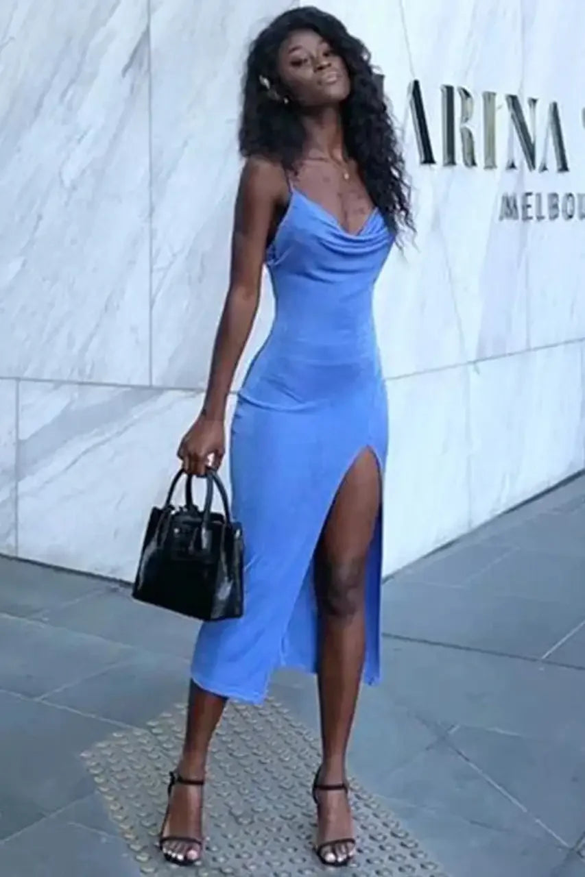 A woman in a blue dress with a thigh-high slit poses confidently, holding a black handbag. She stands on a stone surface in front of a marble wall, radiating elegance.