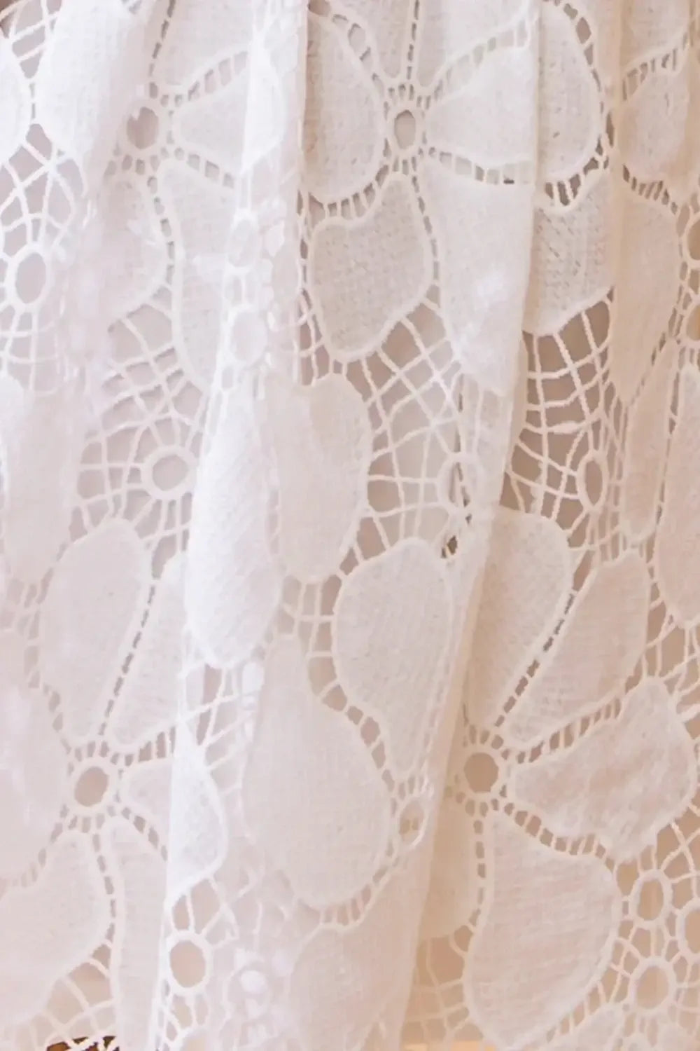 A white lace fabric with intricate floral patterns drapes gracefully. The delicate texture and design suggest a soft, feminine aesthetic, likely used in clothing or home decor.