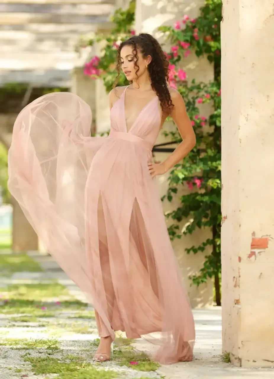 A woman in a flowing, pale pink dress stands in a sunlit outdoor setting, with her arm outstretched, and the fabric billowing gently around her against a backdrop of greenery and stone walls.