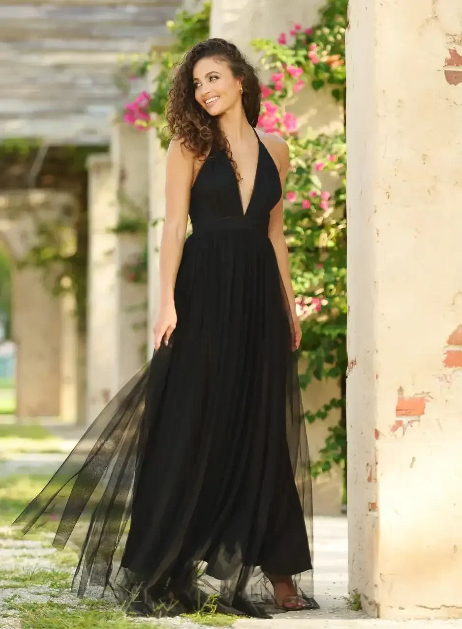 A woman wearing a flowing black dress stands in a garden, smiling. She is surrounded by stone walls adorned with blooming plants, creating a serene, elegant atmosphere.