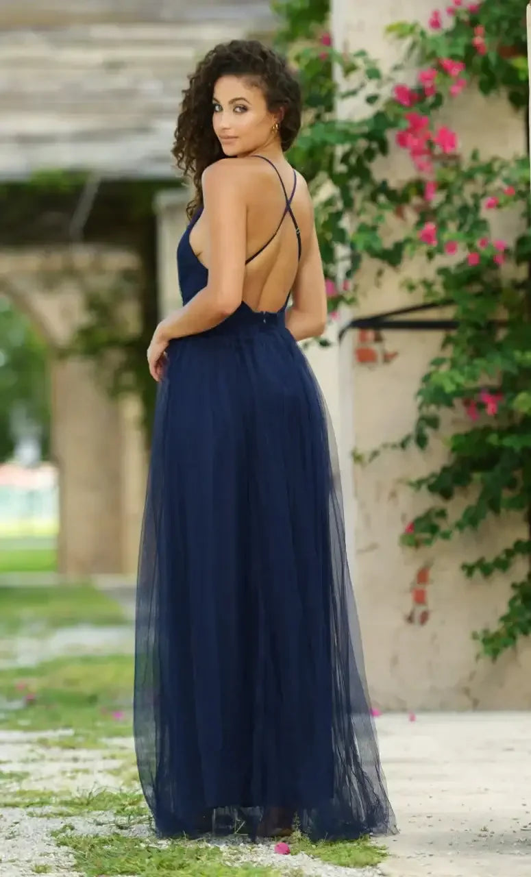 A woman wearing a navy blue, backless gown stands facing away, gently holding her waist. She is in an outdoor setting with greenery and pink flowers in the background.