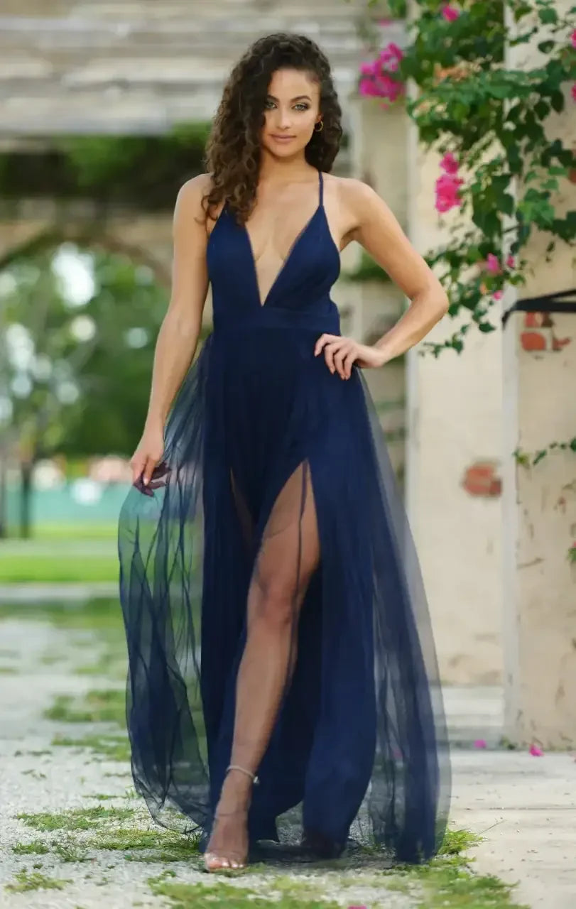 A woman wears a flowing, navy blue gown with a deep V-neck and thigh-high splits, confidently standing in a lush, outdoor setting decorated with pink flowers and greenery.
