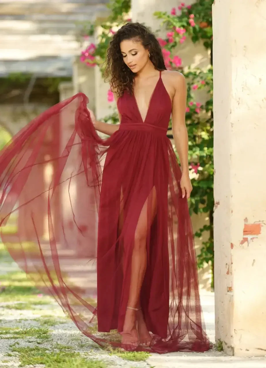 A woman in a flowing burgundy dress is elegantly posing outdoors, gently holding the fabric. The backdrop features sunlit stone walls and vibrant pink flowers, creating a serene, romantic atmosphere.