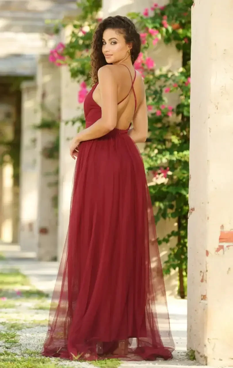 A woman wears a flowing burgundy dress and poses gracefully, looking over her shoulder. She stands in a garden-like setting with greenery and pink flowers framing the background.