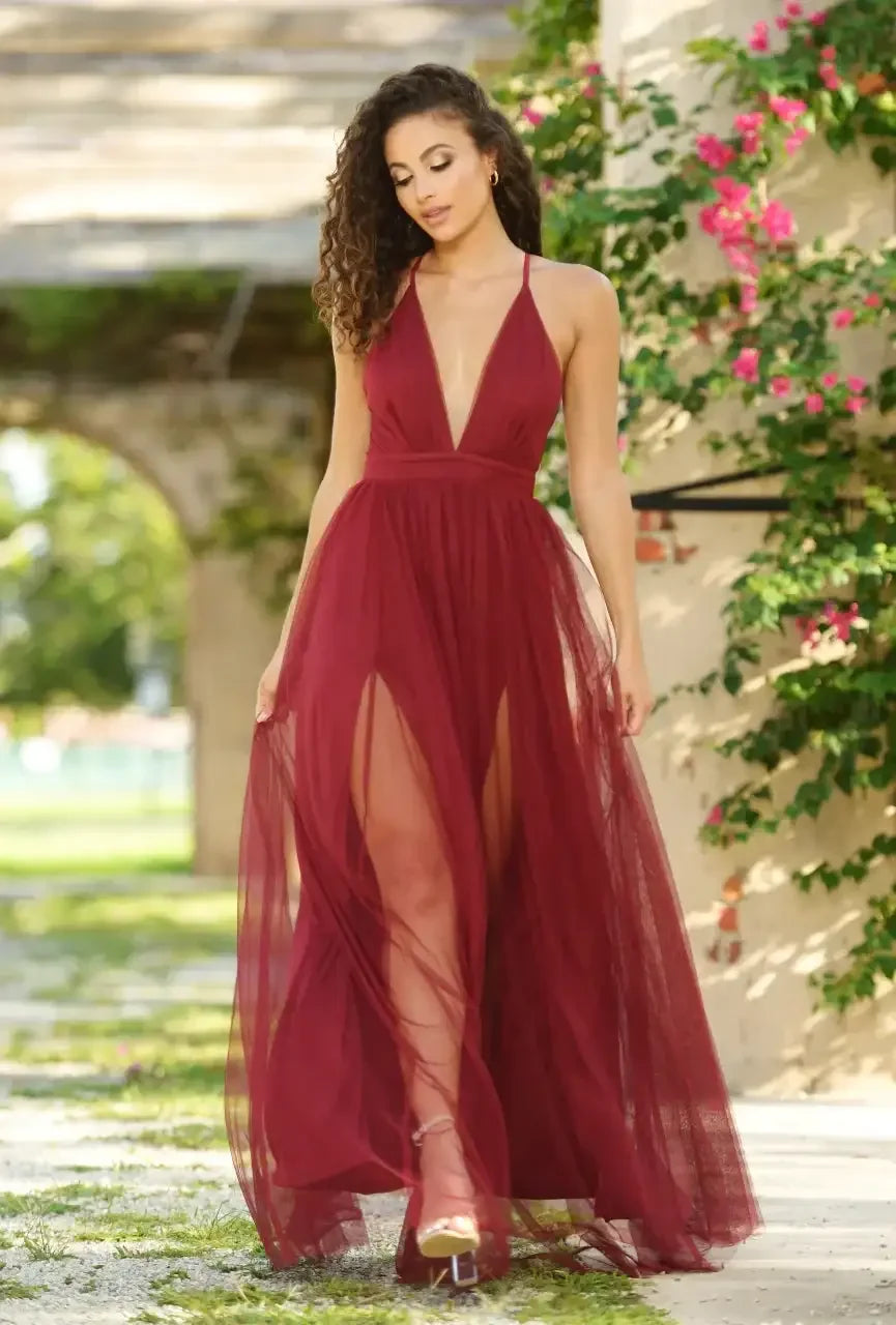 A woman wears a flowing burgundy dress with a deep neckline and high slits, gracefully walking through a sunlit garden adorned with lush greenery and vibrant flowers.