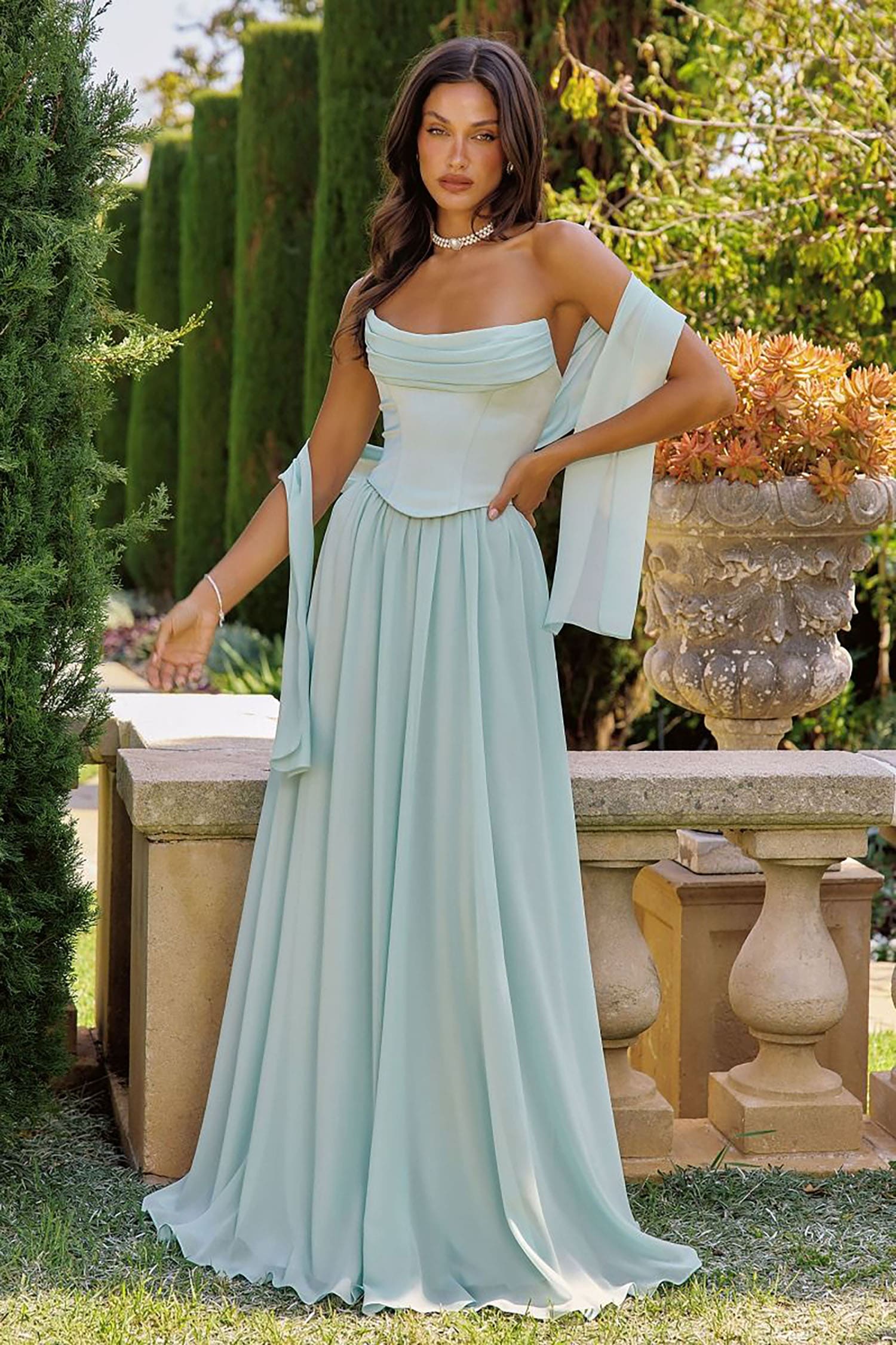 A woman models a floor-length off-the-shoulder gown in a pale mint green or light aqua color, standing outdoors on grass beside a stone bench with hedges and a decorative planter in the background.