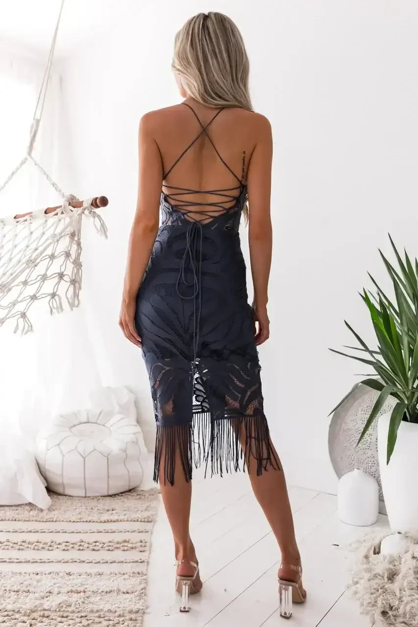 A woman stands with her back turned, wearing a fitted navy dress with fringes. She is in a bright, airy room decorated with plants, a hammock, and soft furnishings.