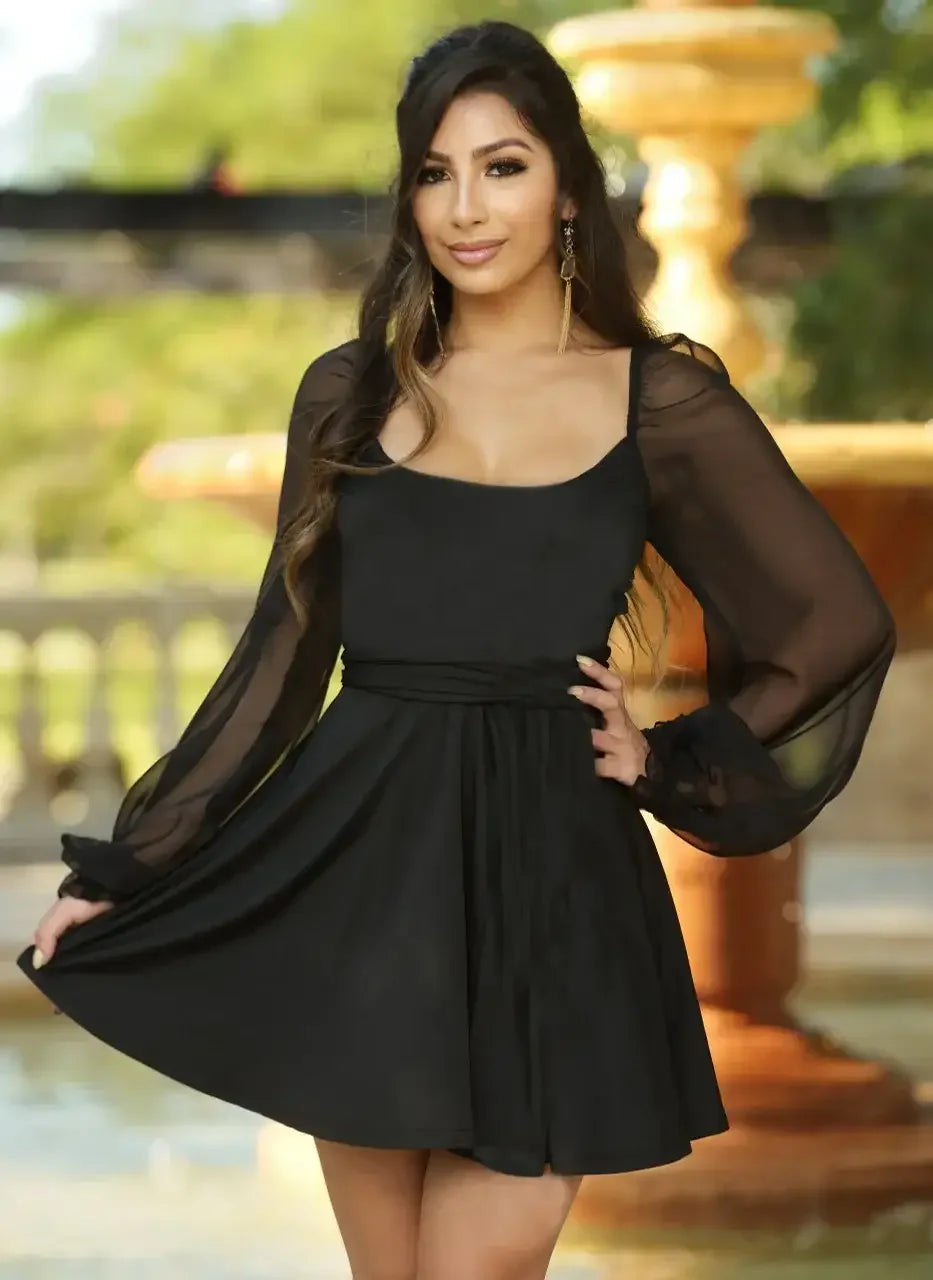 A woman wears a short black dress with sheer long sleeves, smiling confidently. She stands near a decorative fountain surrounded by greenery, creating a vibrant outdoor setting.