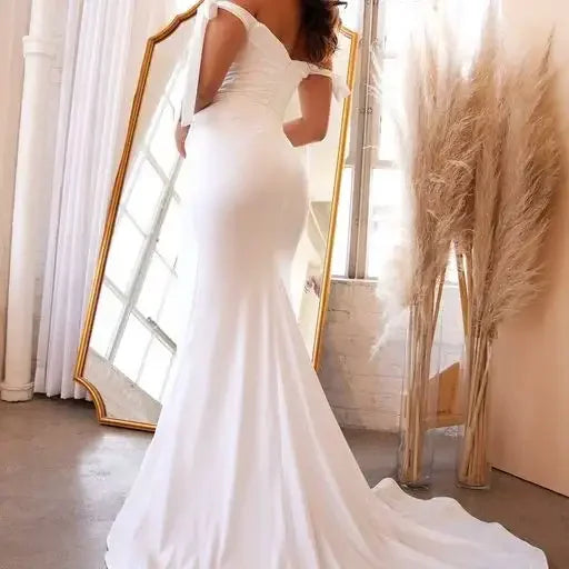 A person in a flowing white dress stands sideways, exposing the garment's form-fitting silhouette. They are in a bright, airy room with large windows and decorative pampas grass nearby.