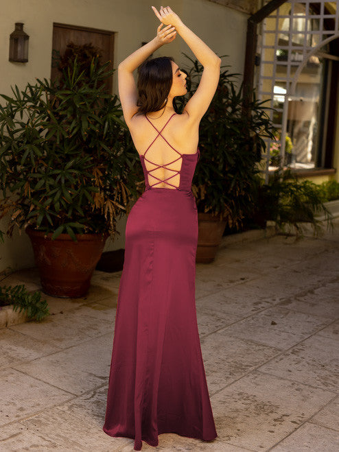 A woman in a burgundy dress with a crisscross back poses gracefully, stretching her arms upward. She stands on a tiled path surrounded by lush potted plants and an elegant entrance.