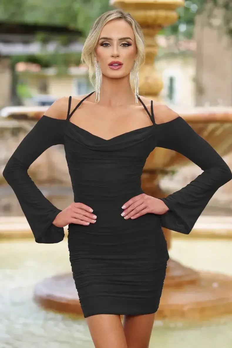 A woman stands confidently in a fitted black dress with long, flared sleeves. She poses with her hands on her hips, set against a decorative stone fountain in a garden.
